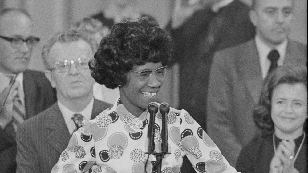 Shirley Chisholm smiling and speaking at a podium, flanked by onlookers in a formal setting.