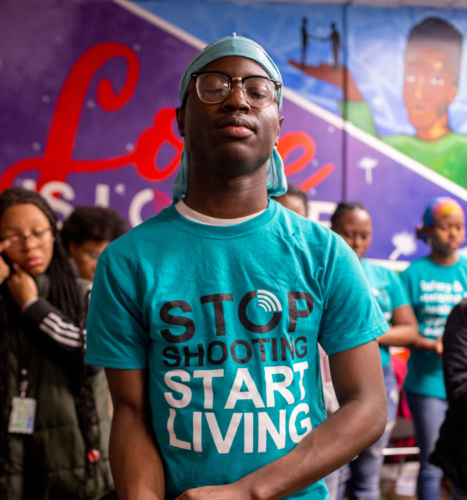 A man stands in the foreground wearing a teal t-shirt with the phrase "stop shooting start living" printed on it, while other individuals appear in the background.