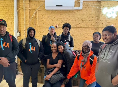 A group of eight individuals posing for a group photo indoors, with most wearing similar black clothing featuring a distinctive logo.