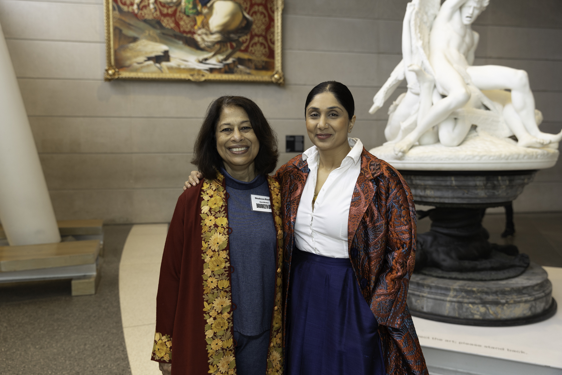 Two women smiling for a photo in an art gallery with paintings and a sculpture in the background.