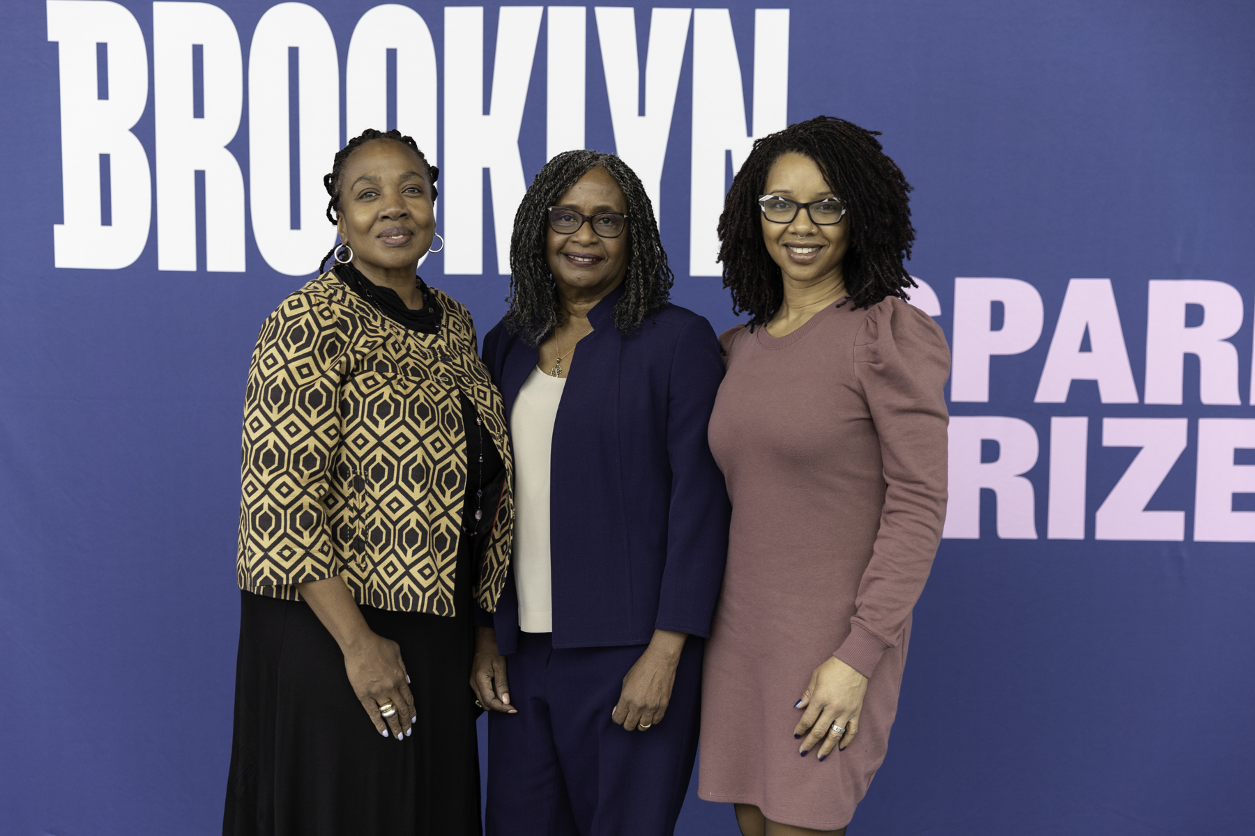 Three women smiling and posing together in front of a blue backdrop with the word "brooklyn.