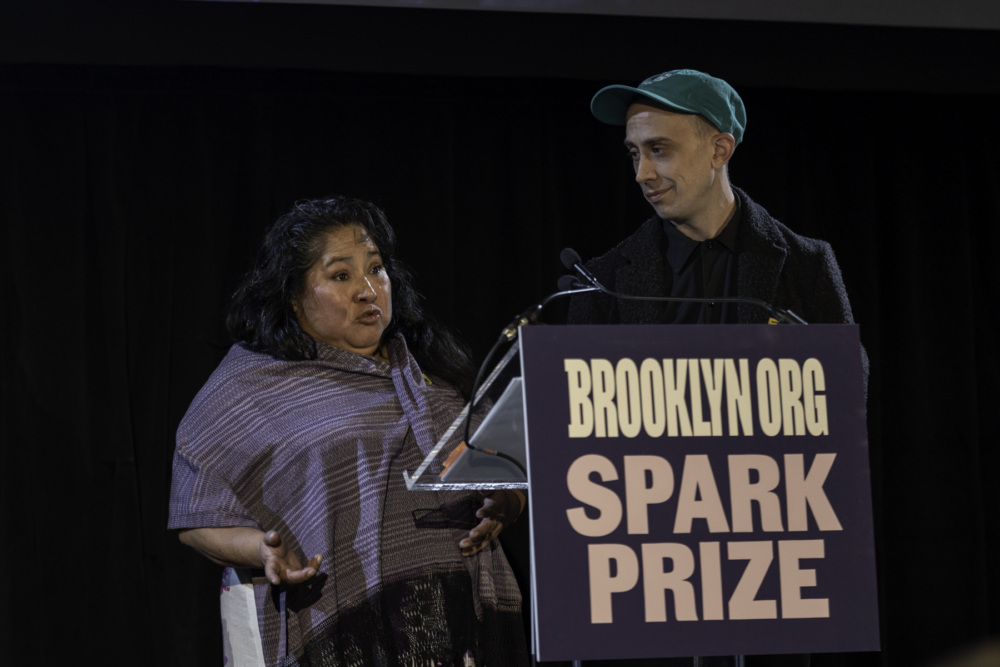 Two individuals at a podium with a microphone, with a sign reading "brooklyn.org spark prize," at an event.