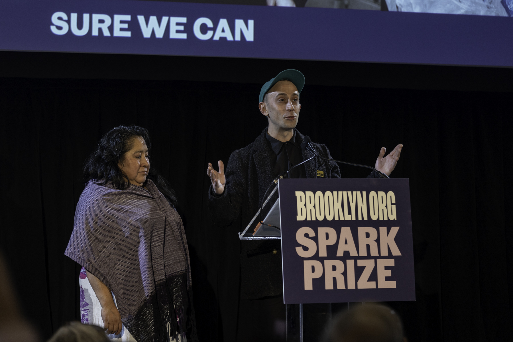 A speaker addresses an audience at a podium with the "brooklyn.org spark prize" sign, accompanied by a person wearing a shawl to their left.
