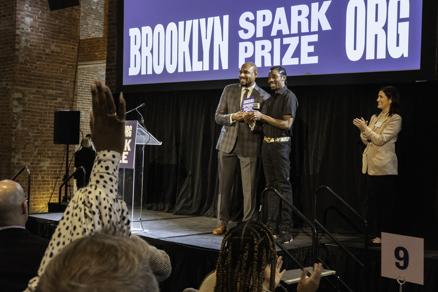 Two individuals holding an award on stage at a brooklyn spark prize event while an audience member raises their hand in applause.