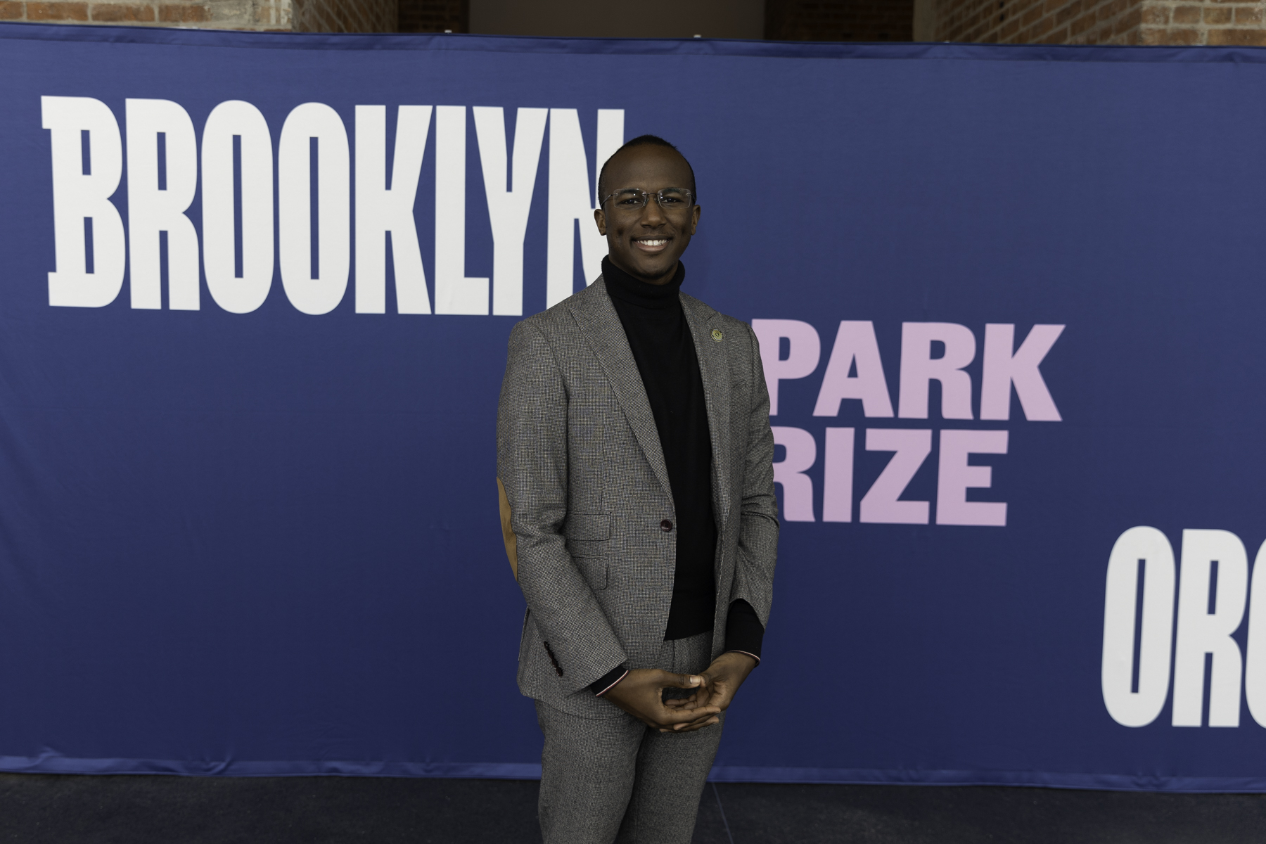 Man in a grey suit and black turtleneck posing in front of a "brooklyn park prize" banner.