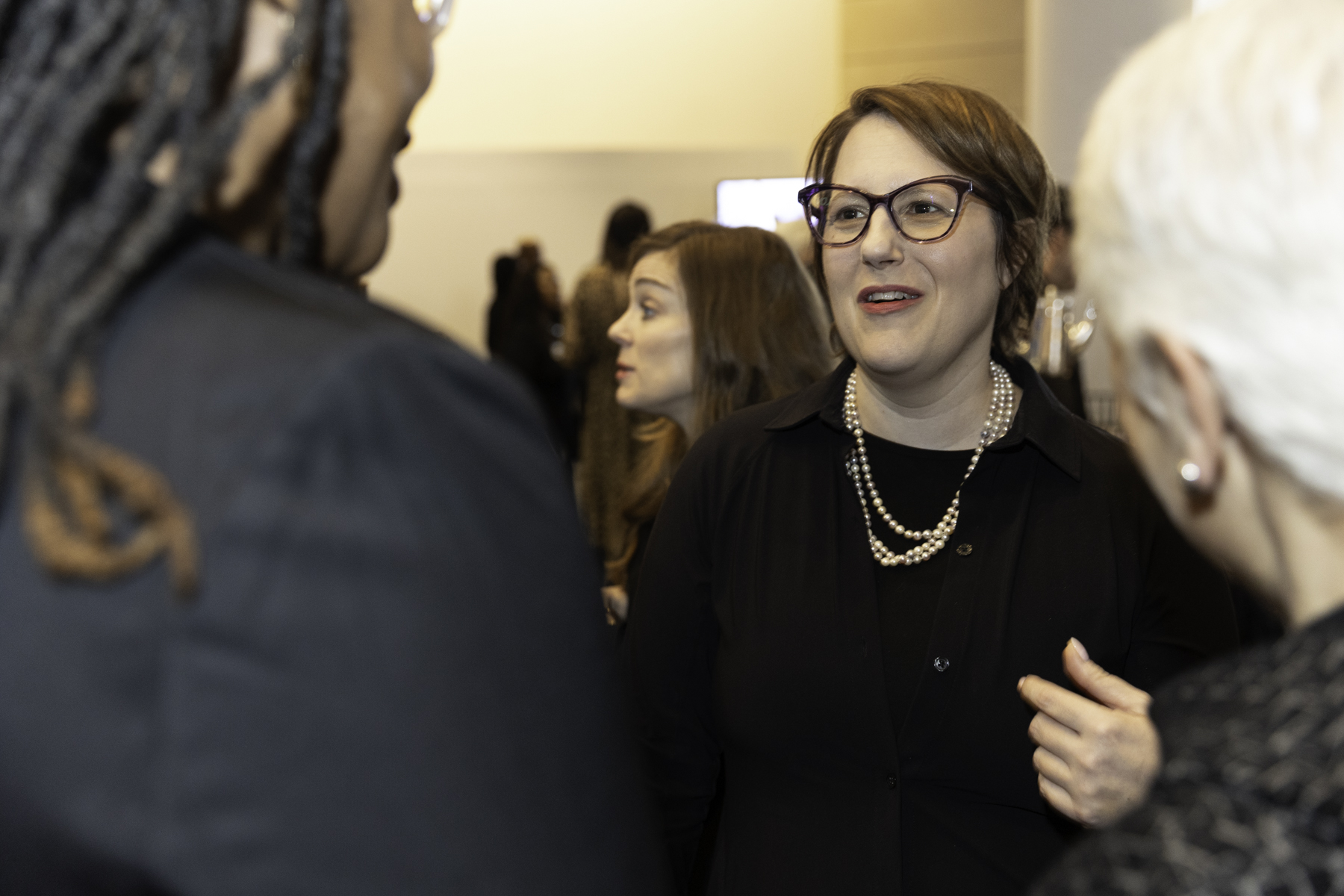A woman in glasses and a black outfit with a pearl necklace is conversing with others at a social gathering.