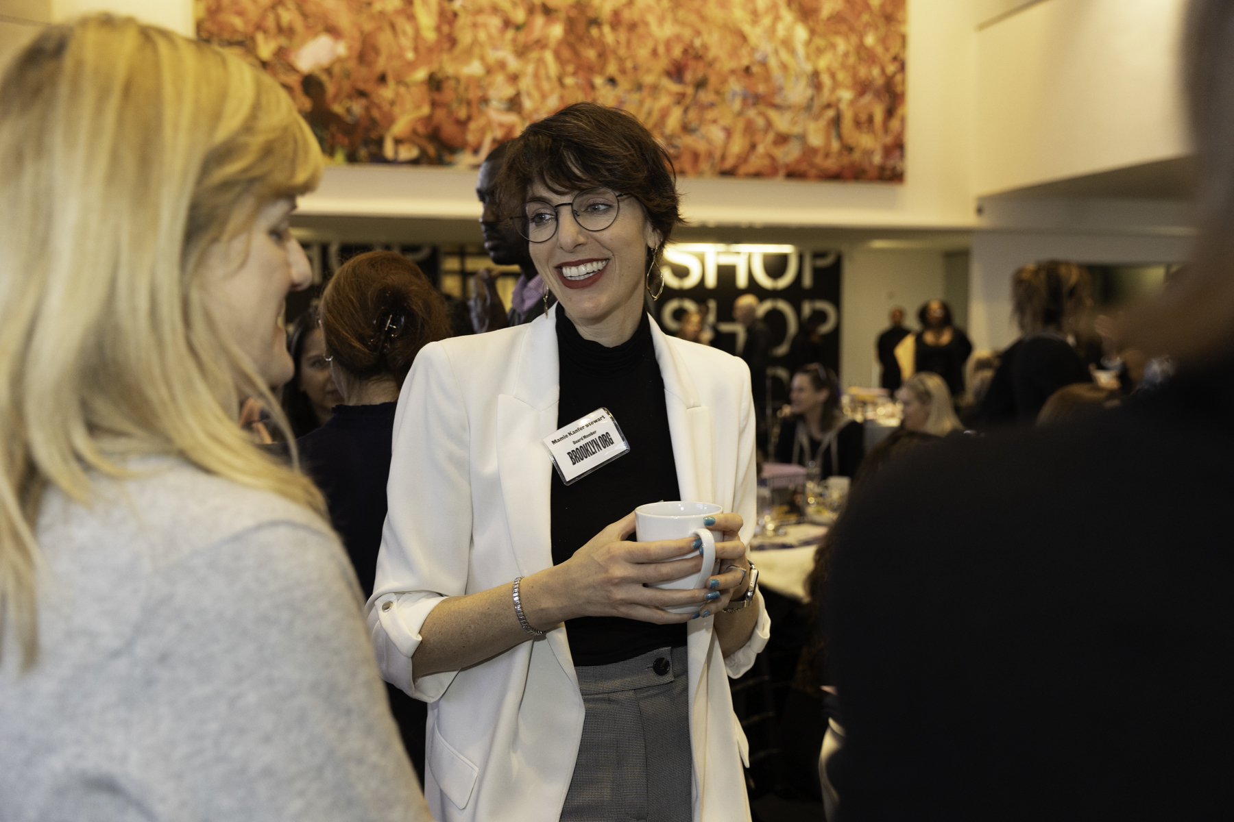 A woman with a name tag holds a coffee cup while smiling and conversing with others at a social event.