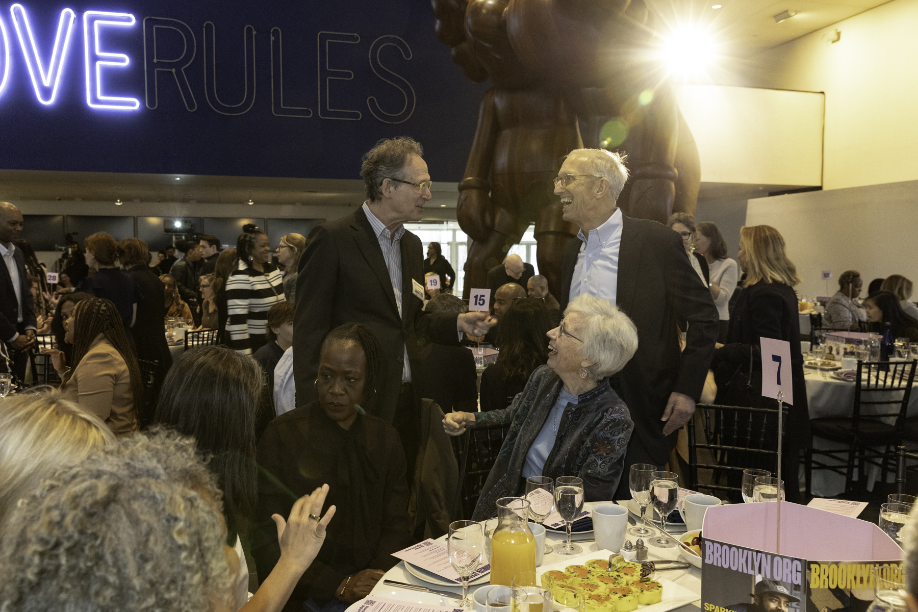 Two men engaged in a standing conversation at a crowded event with seated guests and a large sculpture in the background.