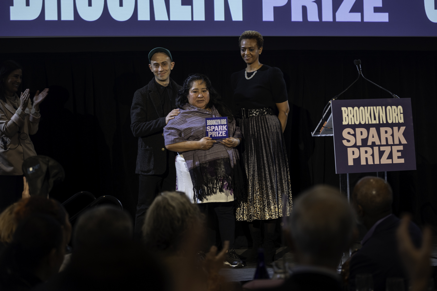 Recipient holding an award at a podium during a brooklyn community foundation event with two presenters standing by.