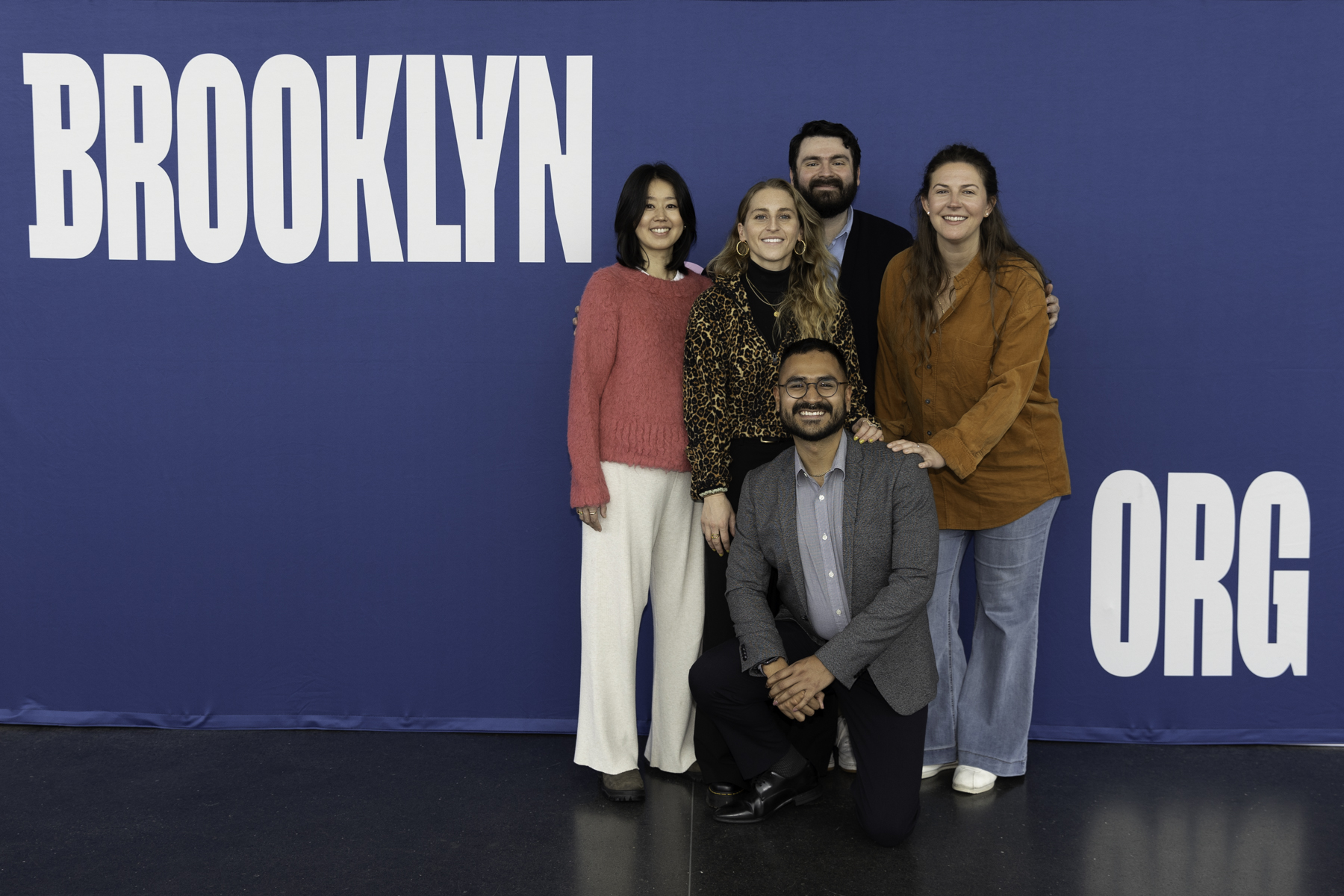 Five individuals posing for a group photo in front of a blue backdrop with the word "brooklyn" displayed prominently.