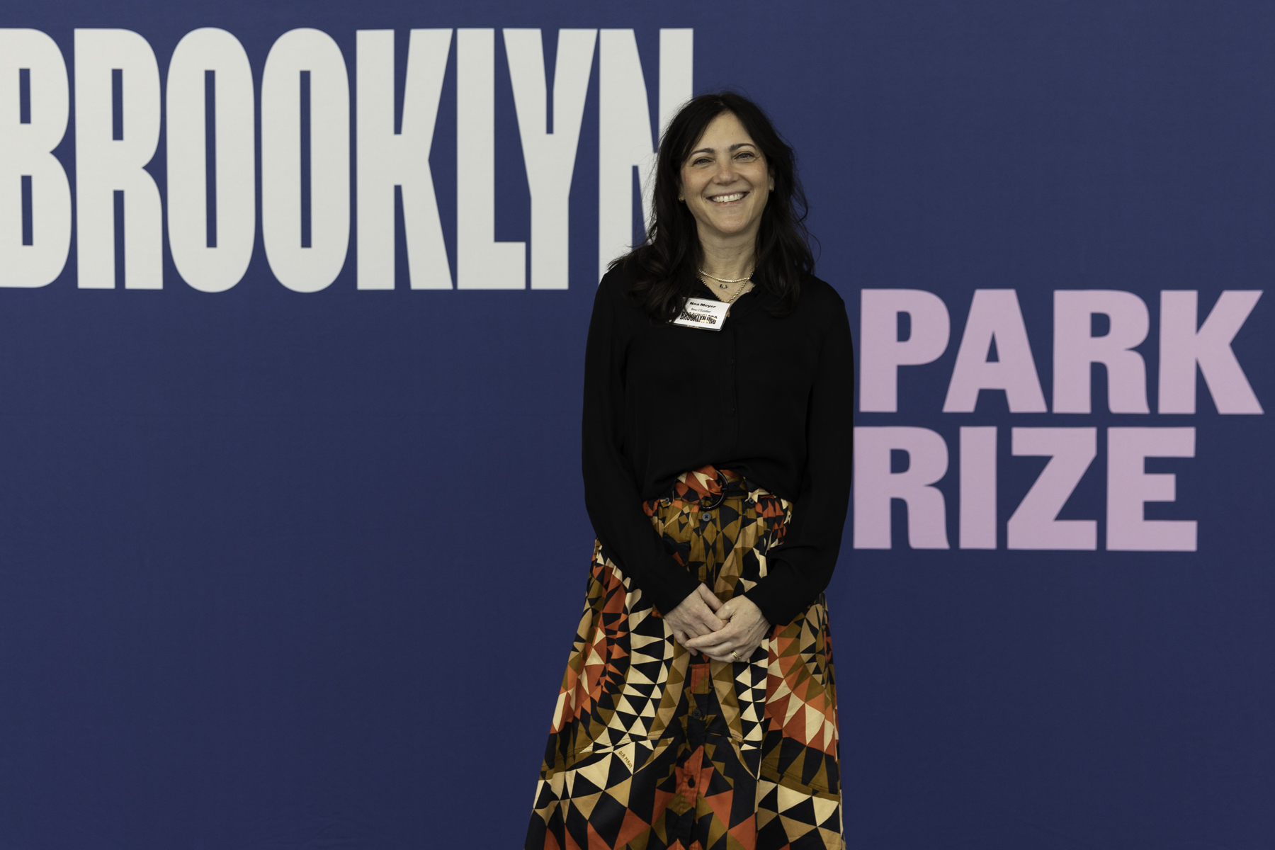 Woman standing in front of a "brooklyn book prize" backdrop with a name tag, smiling for the camera.