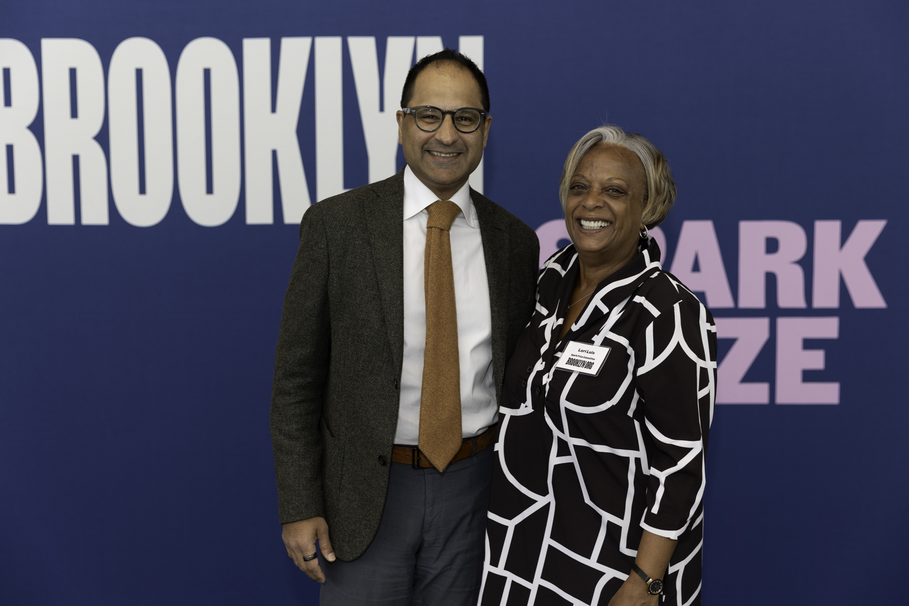 Two individuals smiling for a photo at an event with a backdrop displaying "brooklyn.