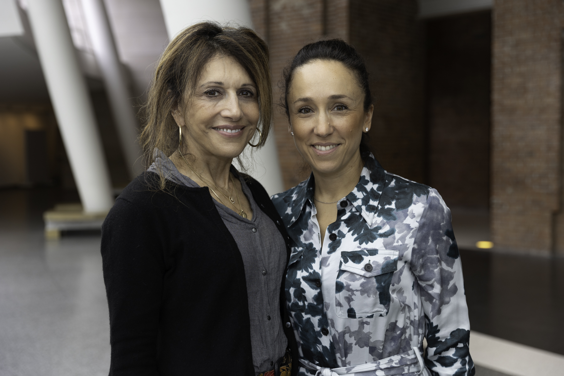 Two women posing for a photograph indoors, smiling at the camera, with one wearing a gray jacket and the other in a camouflaged pattern shirt.
