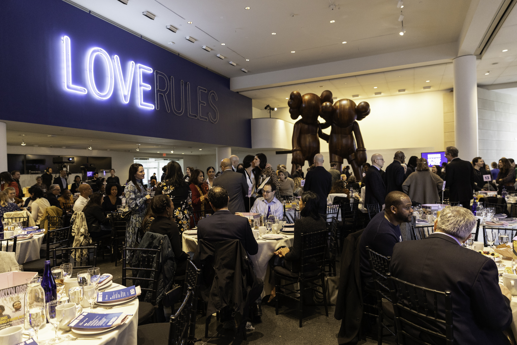 Guests mingling at a formal event with tables set for dinner and a "love rules" neon sign in the background.