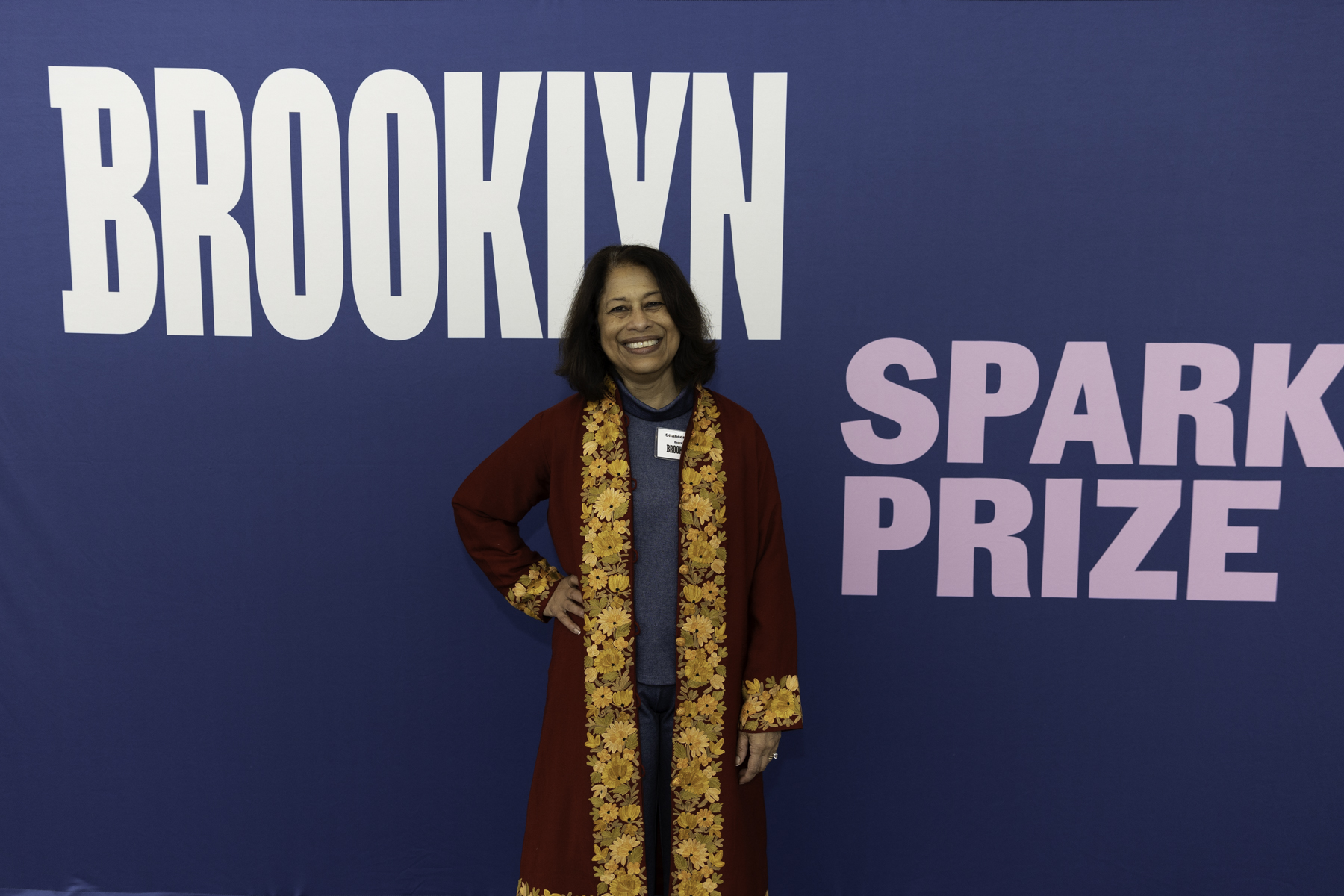 Woman in patterned dress stands smiling before a backdrop with 'brooklyn spark prize' text.