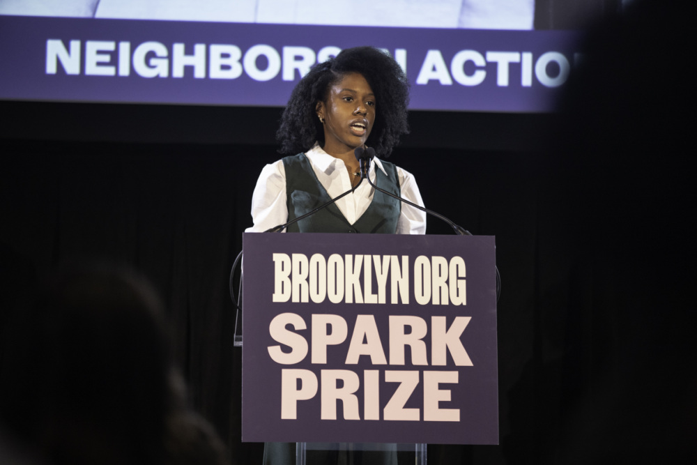 A woman speaking at a podium with a "brooklyn.org spark prize" banner during an event.