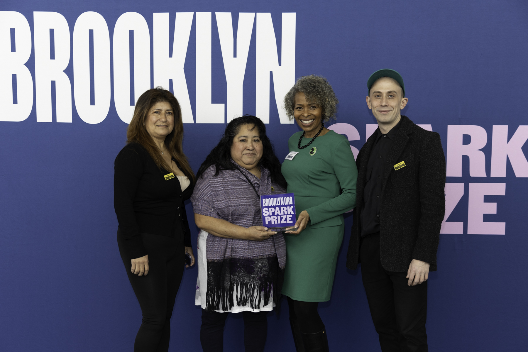 Four individuals posing together in front of a sign that reads "brooklyn" with one holding an award titled "spark prize.