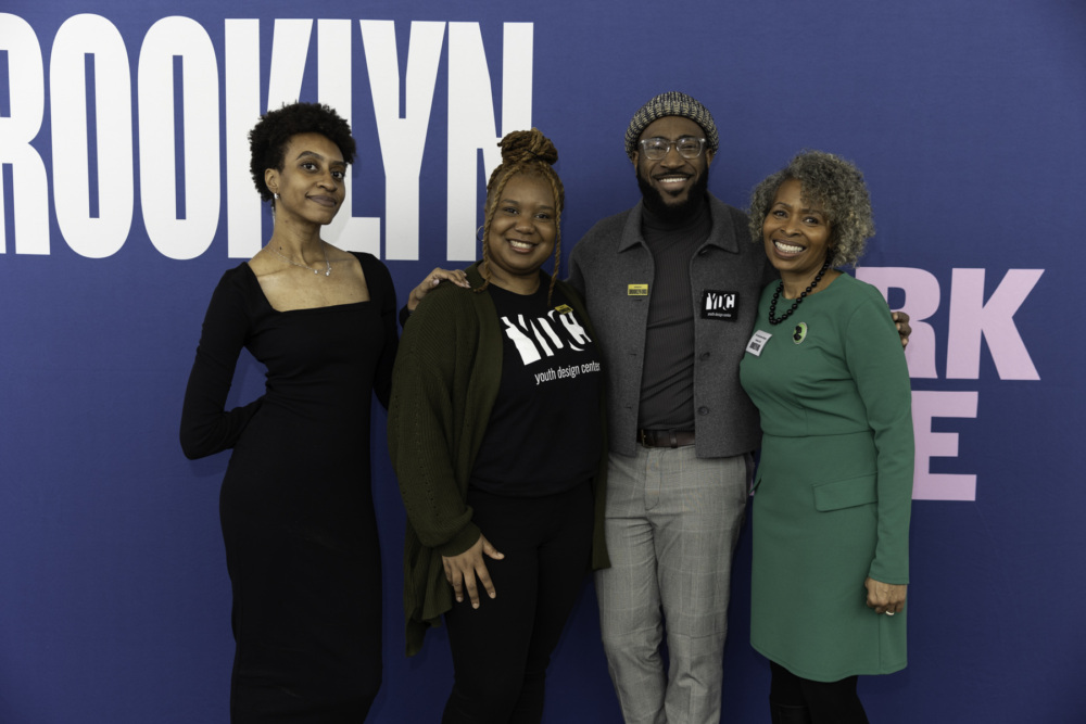 Four individuals posing together in front of a blue backdrop with the word "brooklyn" displayed.