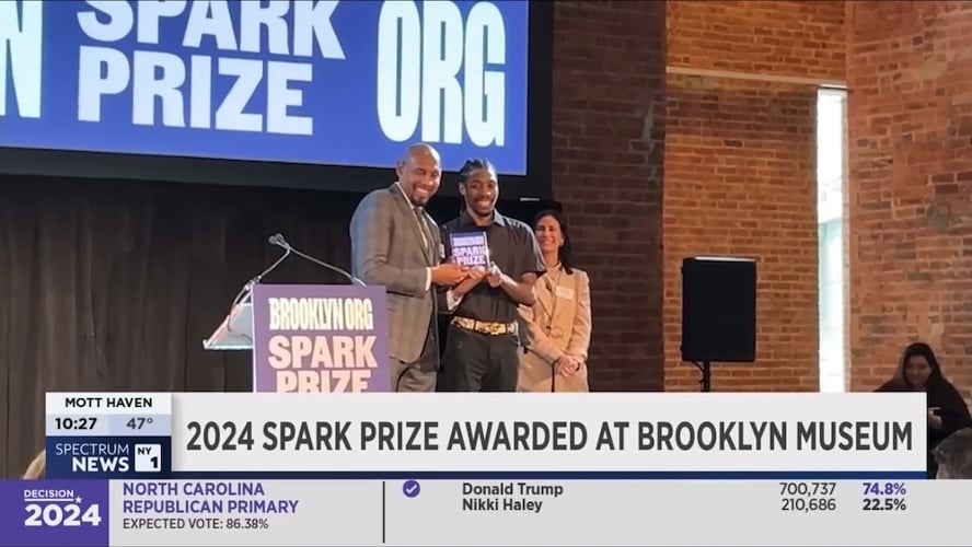 Two individuals receiving an award at the 2024 spark prize event at brooklyn museum, as reported by a news channel with election results displayed at the bottom of the screen.
