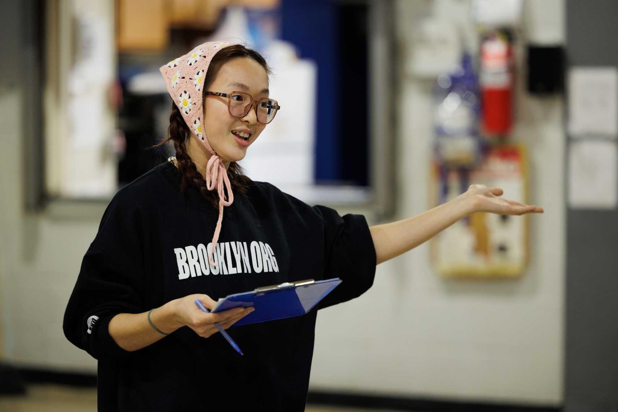 A woman wearing glasses and a pink headscarf speaks and gestures in a workshop setting while holding a clipboard.
