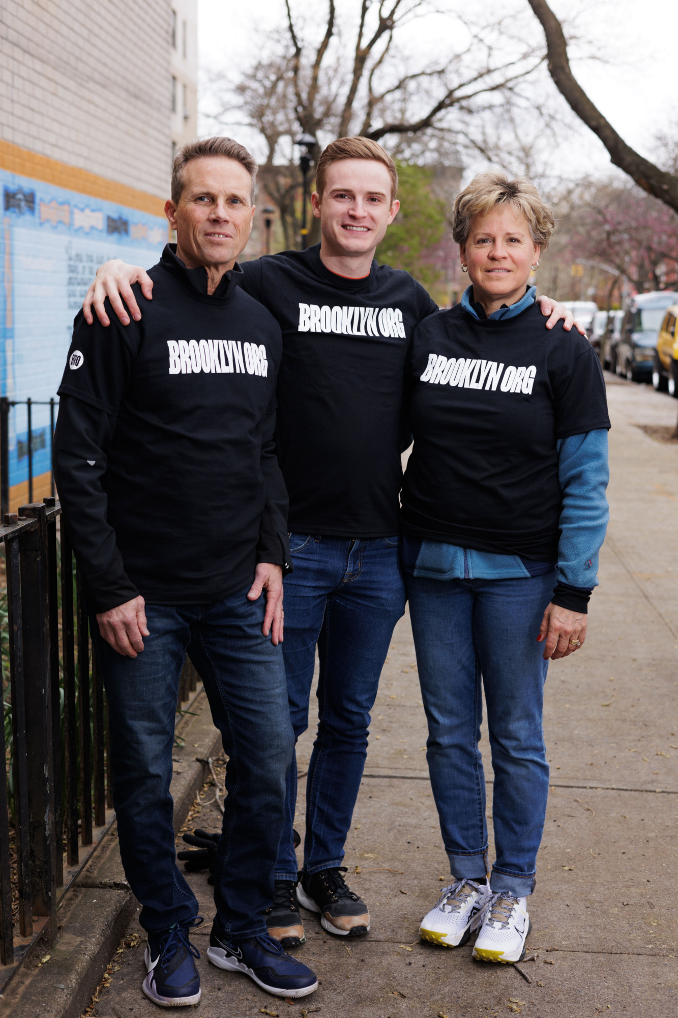 Three people standing on a sidewalk, wearing black "brooklyn" hoodies and jeans, smiling at the camera.