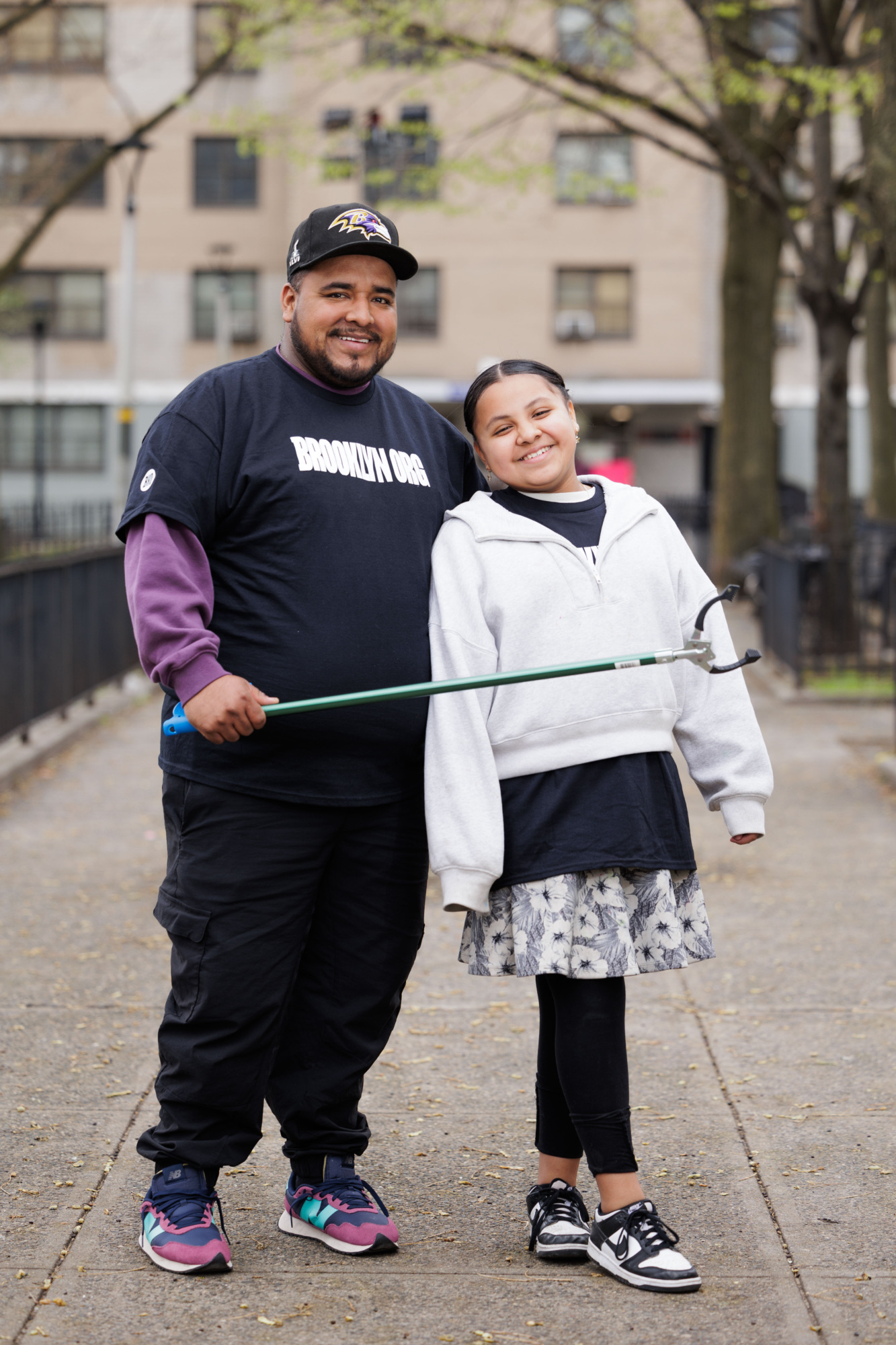 A man and a young girl standing together in a park, smiling at the camera, with the man holding a green stick.