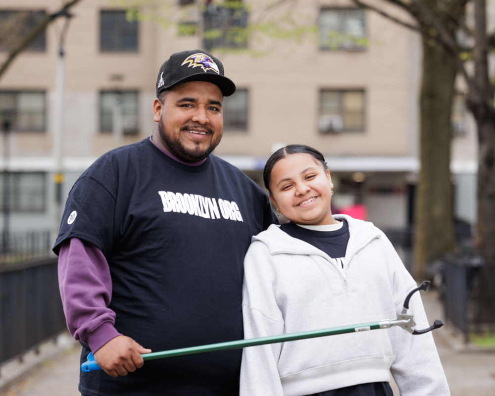 A man and a young girl standing together in a park, smiling at the camera, with the man holding a green stick.