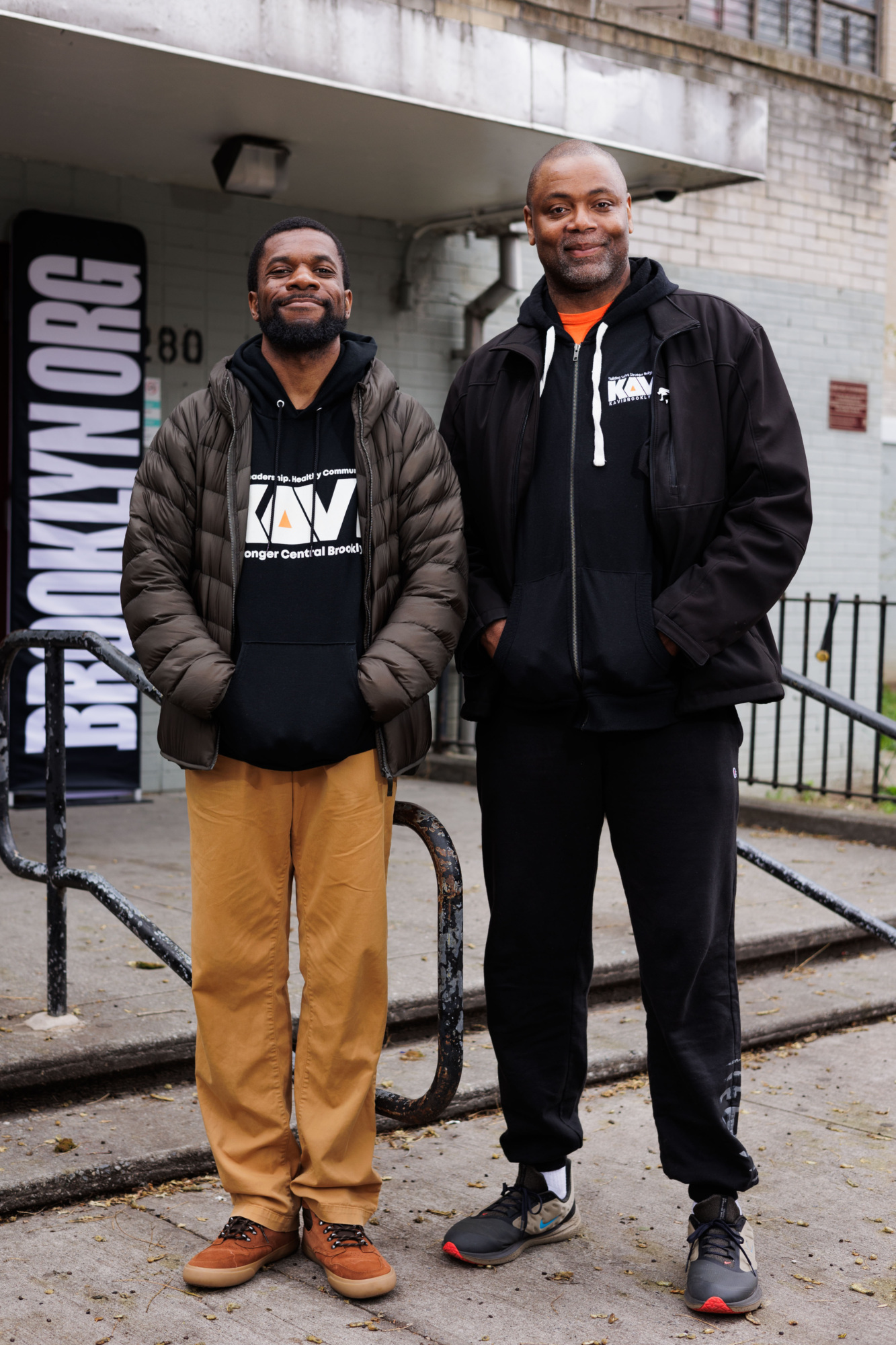 Two smiling men standing in front of a building with a sign that reads "skywork." one wears a black logo hoodie and khakis, the other a black jacket and jeans.