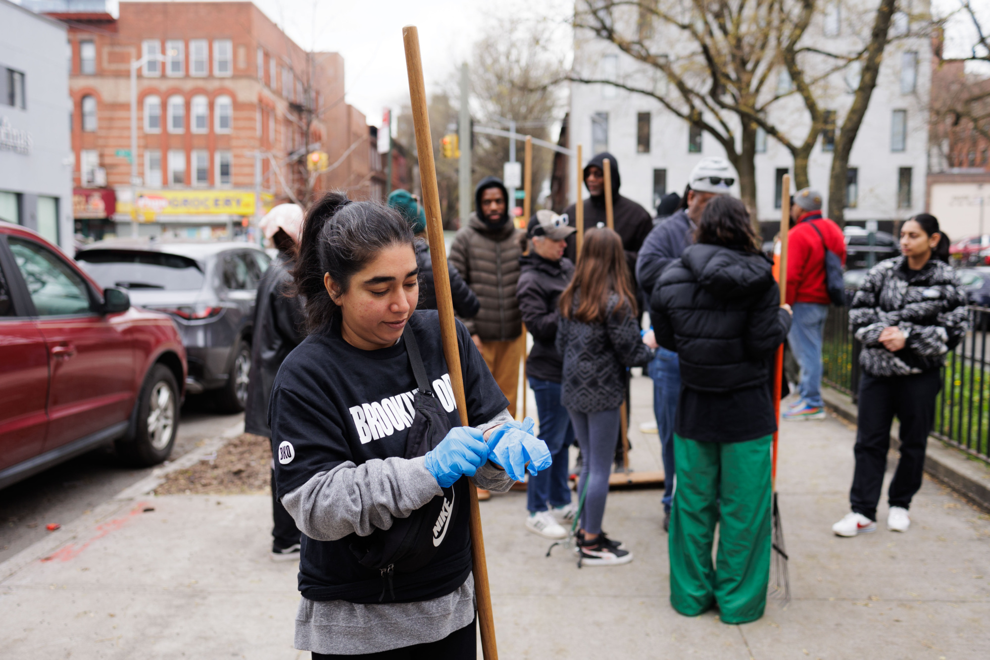 A woman wearing gloves and holding a wooden stick at a community cleaning event on a city street, with other participants in the background.