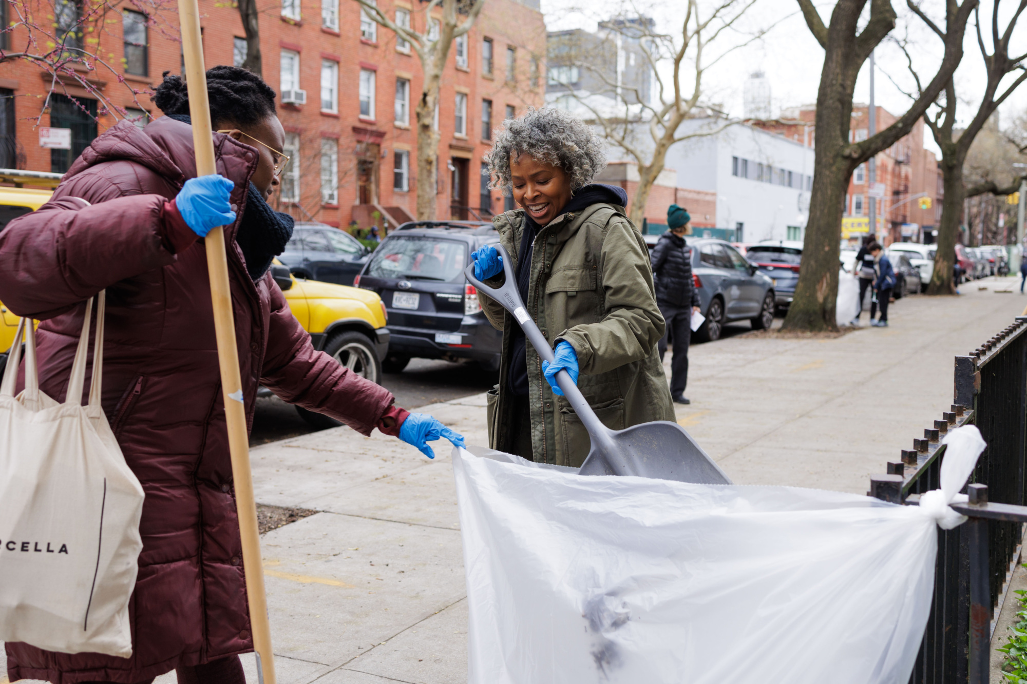 Two people with gloves on smiling as they collect litter with a trash grabber and a bag on a city street.