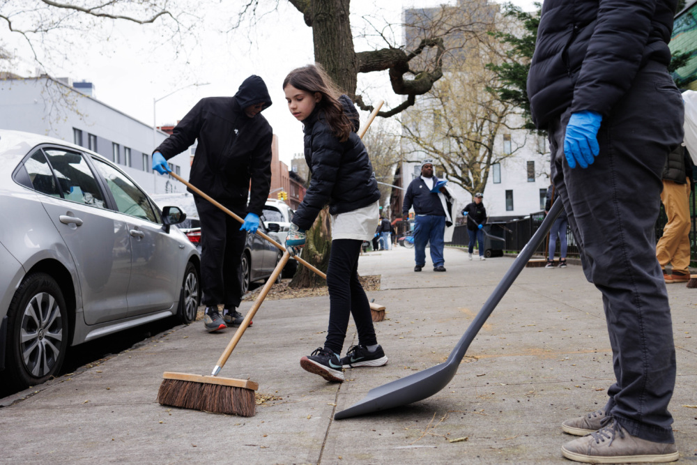 A young girl and a man sweep leaves on a city street, both wearing gloves, with other volunteers and parked cars in the background.