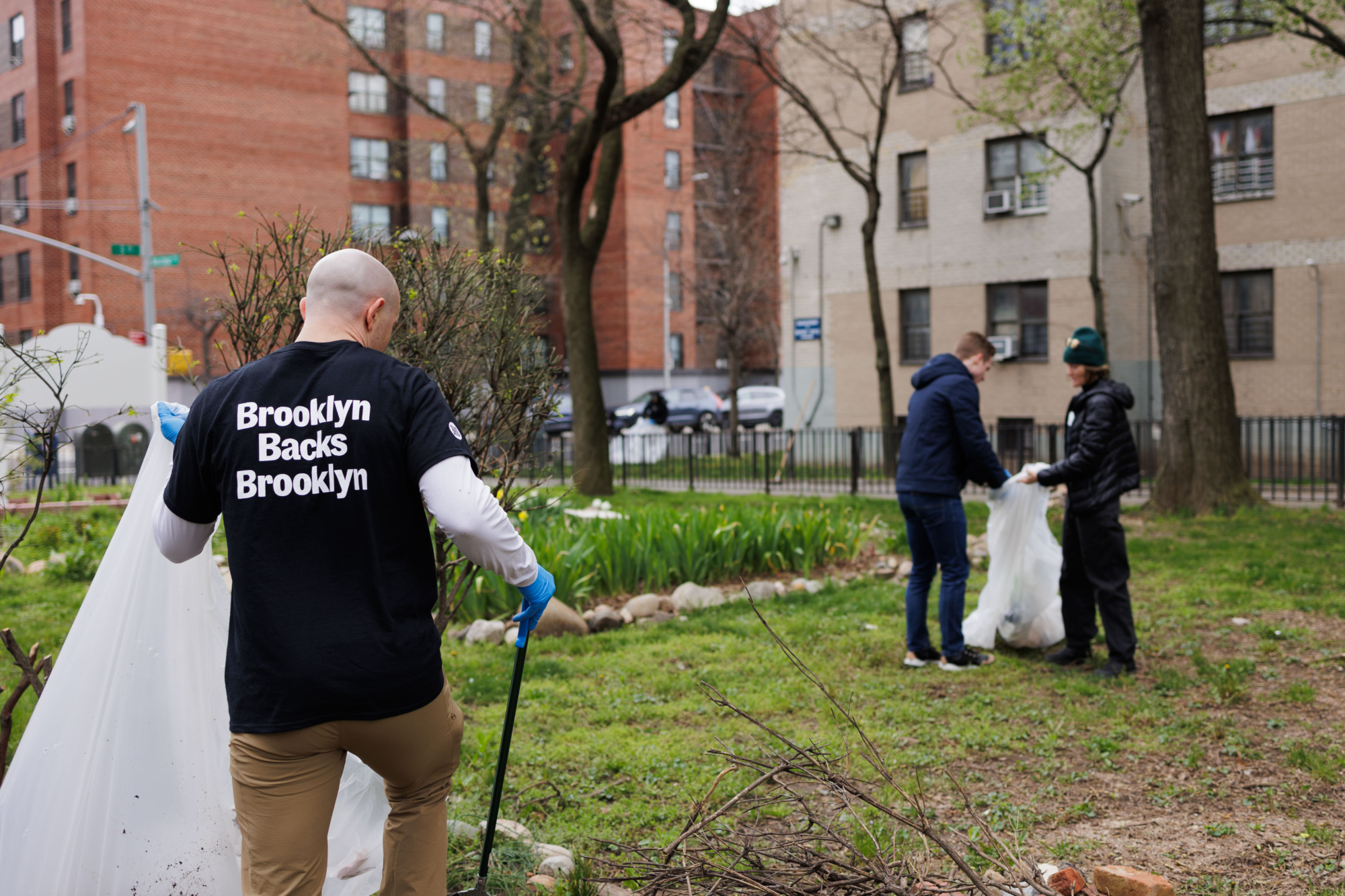 A person in a "brooklyn backs brooklyn" shirt participates in a community clean-up, picking up litter in an urban park with others.