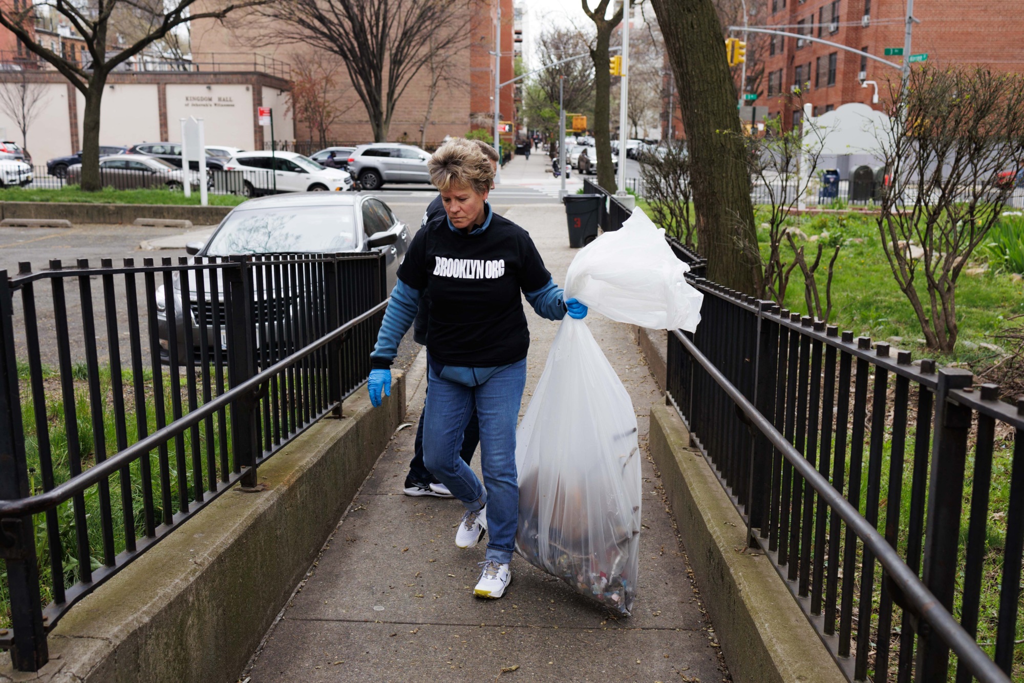 A woman wearing a sweatshirt and gloves carries a large clear trash bag down a small stairway in a city setting.