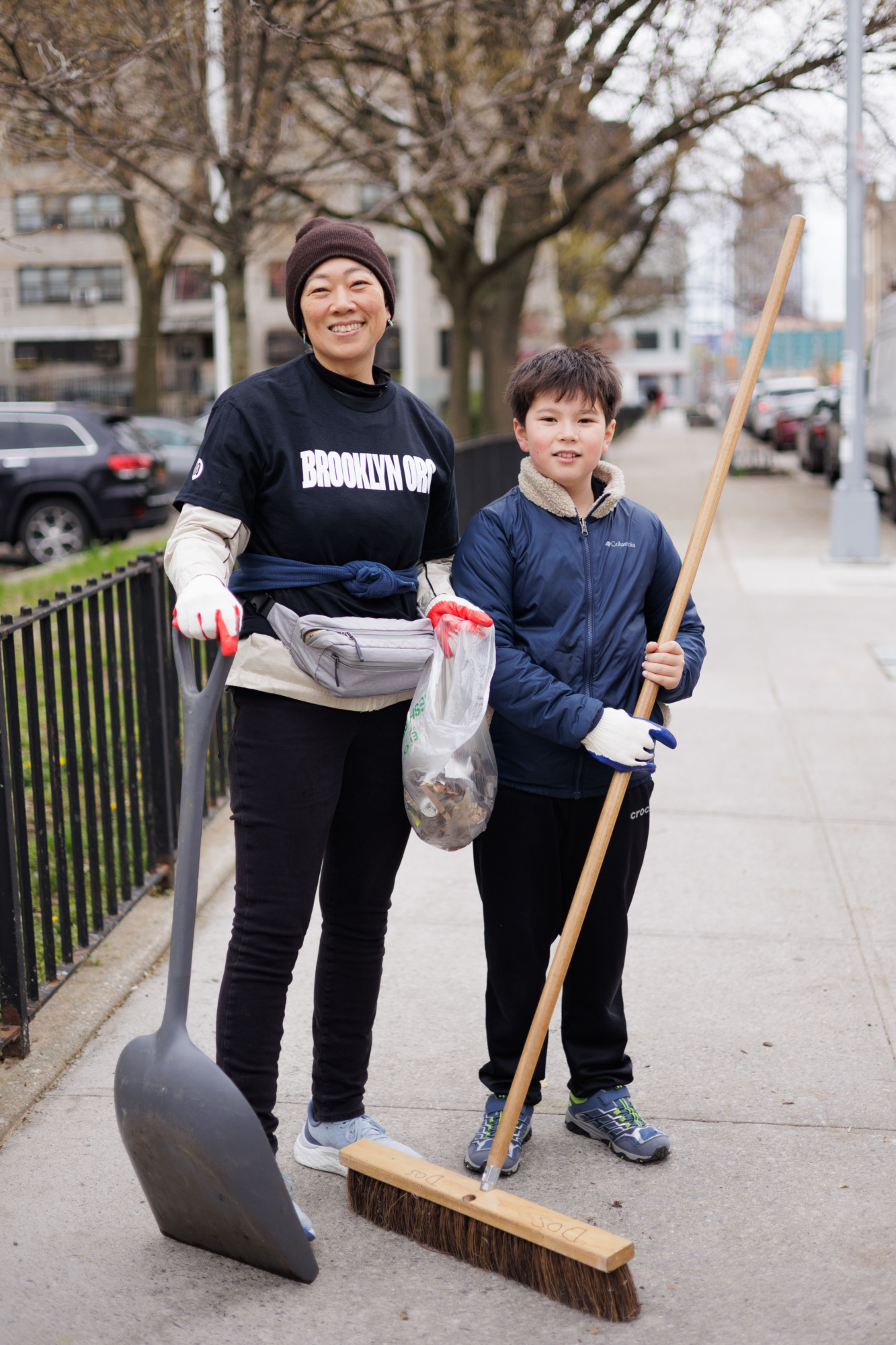 A woman and a young boy, both smiling, hold cleaning tools while participating in a community clean-up event in brooklyn.