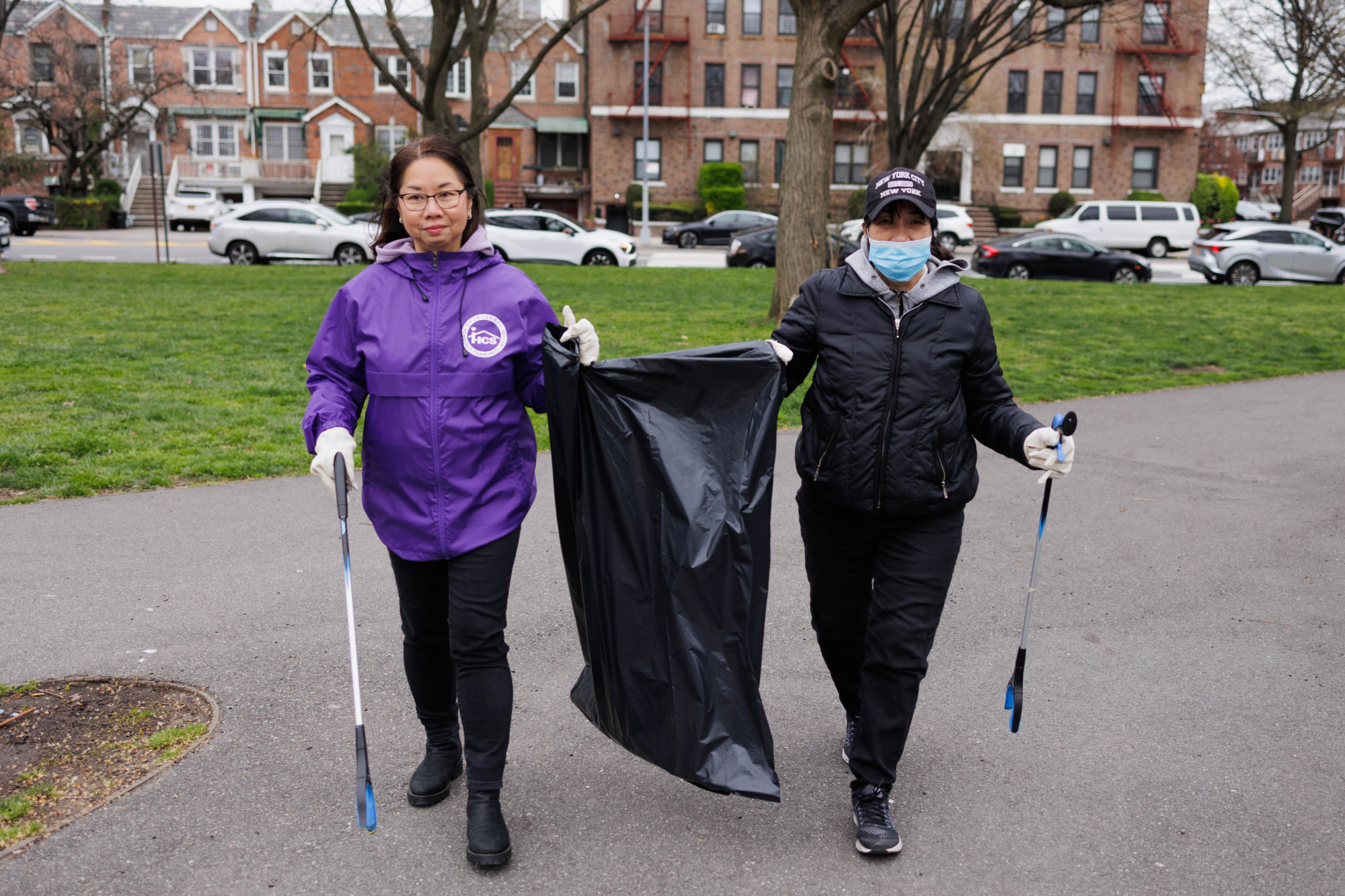 Two people wearing masks and gloves hold trash bags and litter-picking tools in a park, with cars and houses in the background.