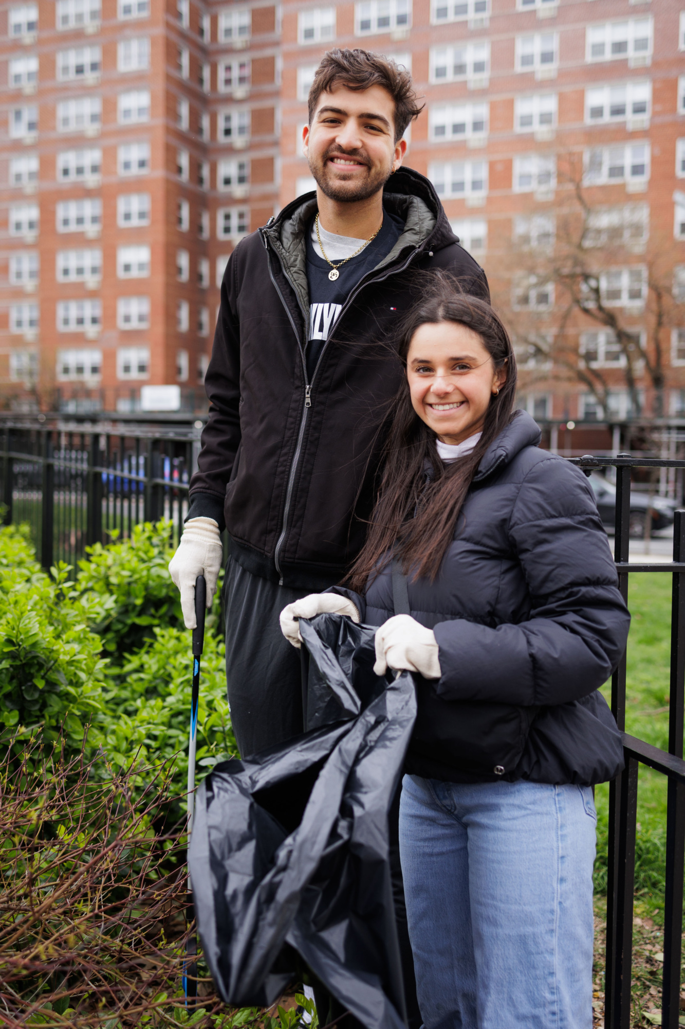 Two volunteers smiling at the camera while holding a trash bag during a community clean-up event in an urban park.
