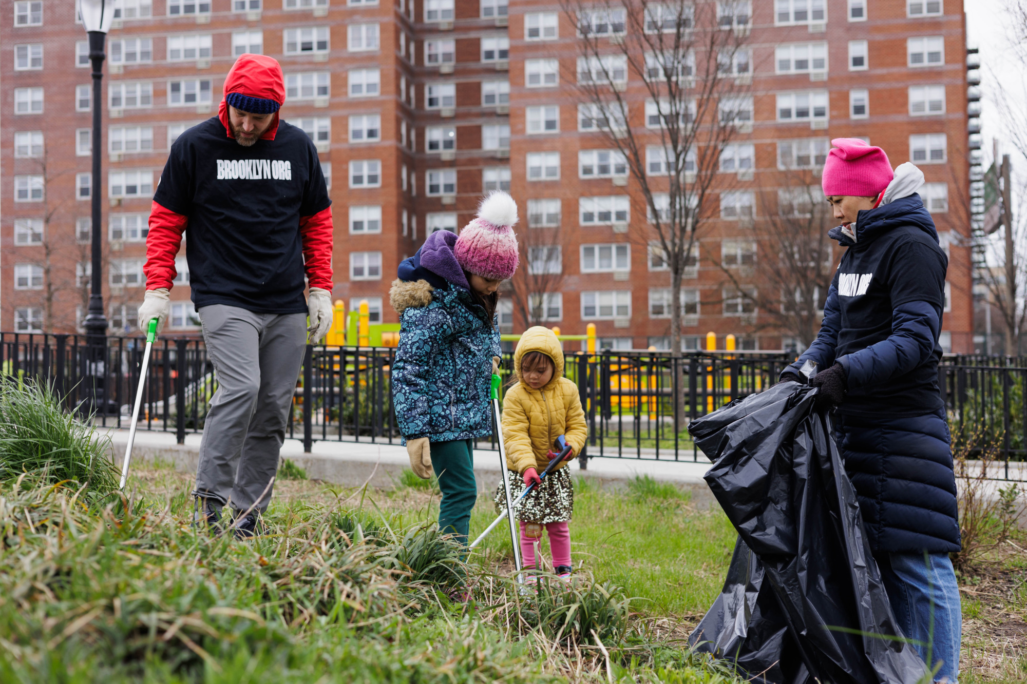 Family volunteers cleaning a park during winter, wearing jackets and hats, with city buildings in the background.