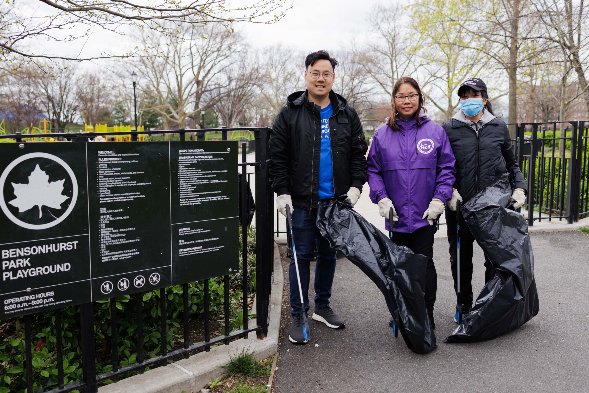 Three volunteers holding garbage bags in a park cleanup event, standing beside a sign for bensonhurst park playground.
