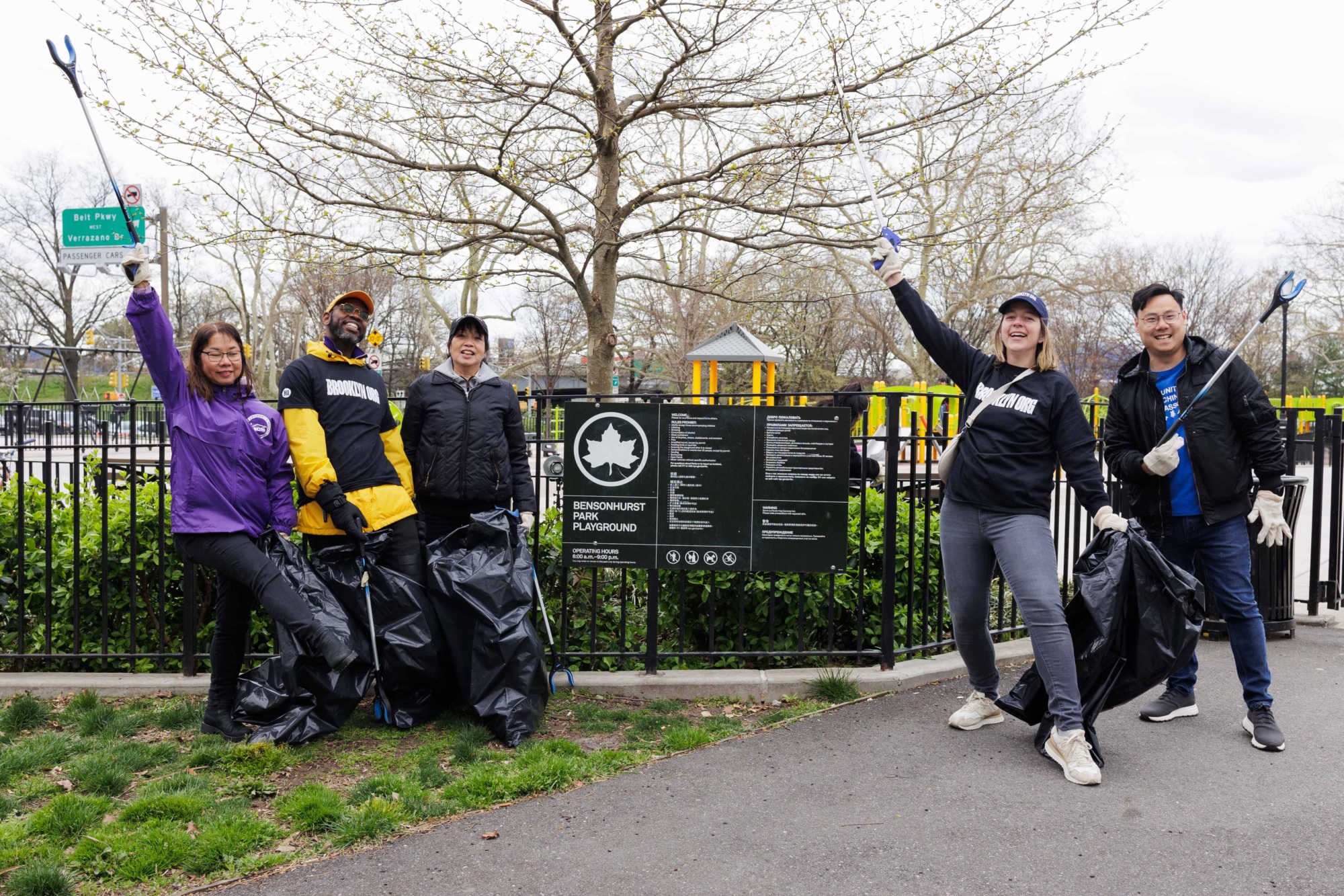 Five volunteers holding trash bags, smiling and celebrating during a clean-up event in a park, with trees and a cloudy sky in the background.