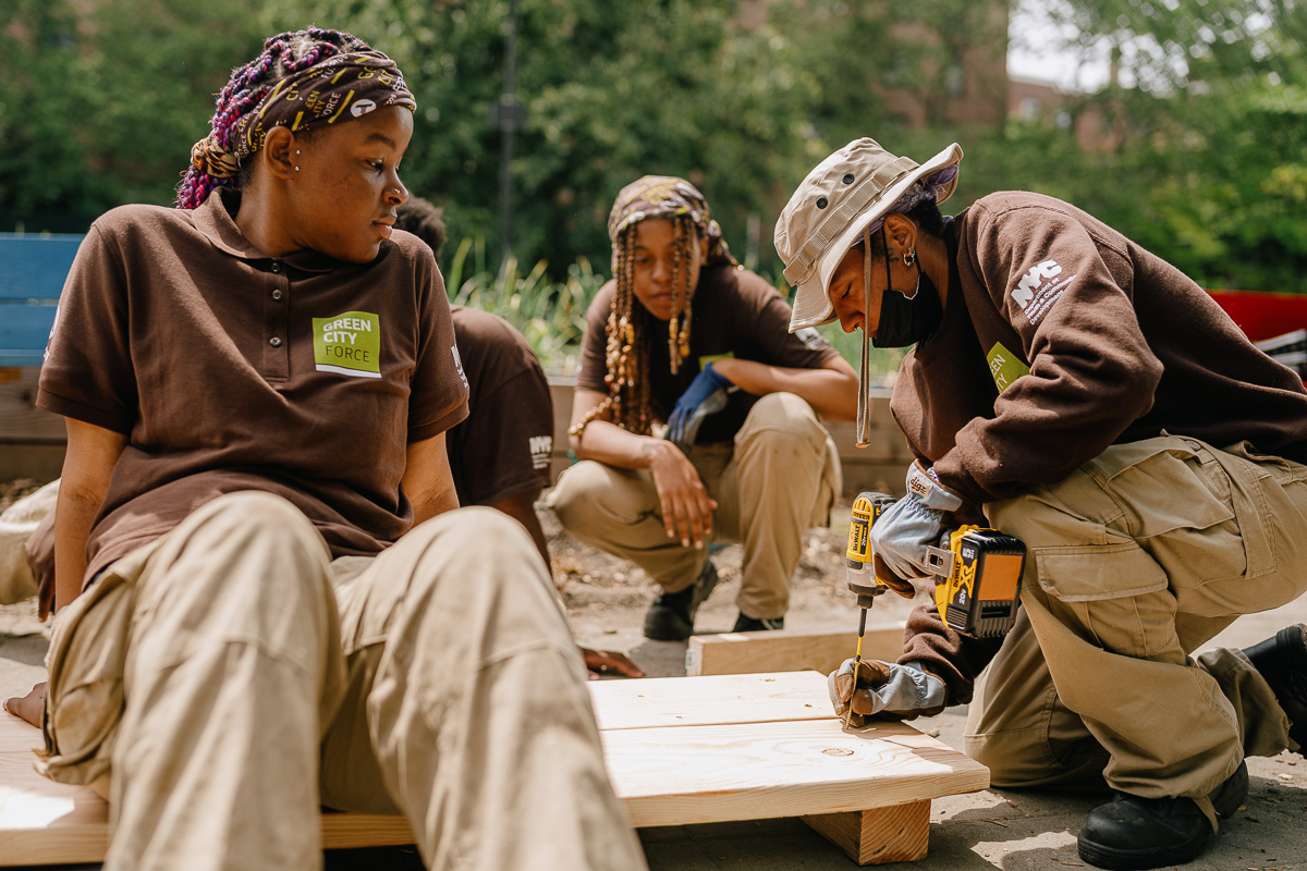 Three individuals in work attire engaged in carpentry, with one using a drill on a wooden plank.