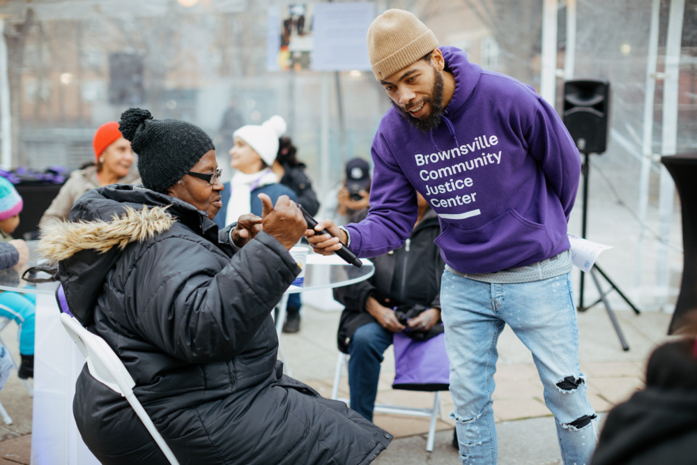 A man in a purple "brownsville community justice center" sweatshirt joyfully arm wrestling with an older woman at an outdoor community event.