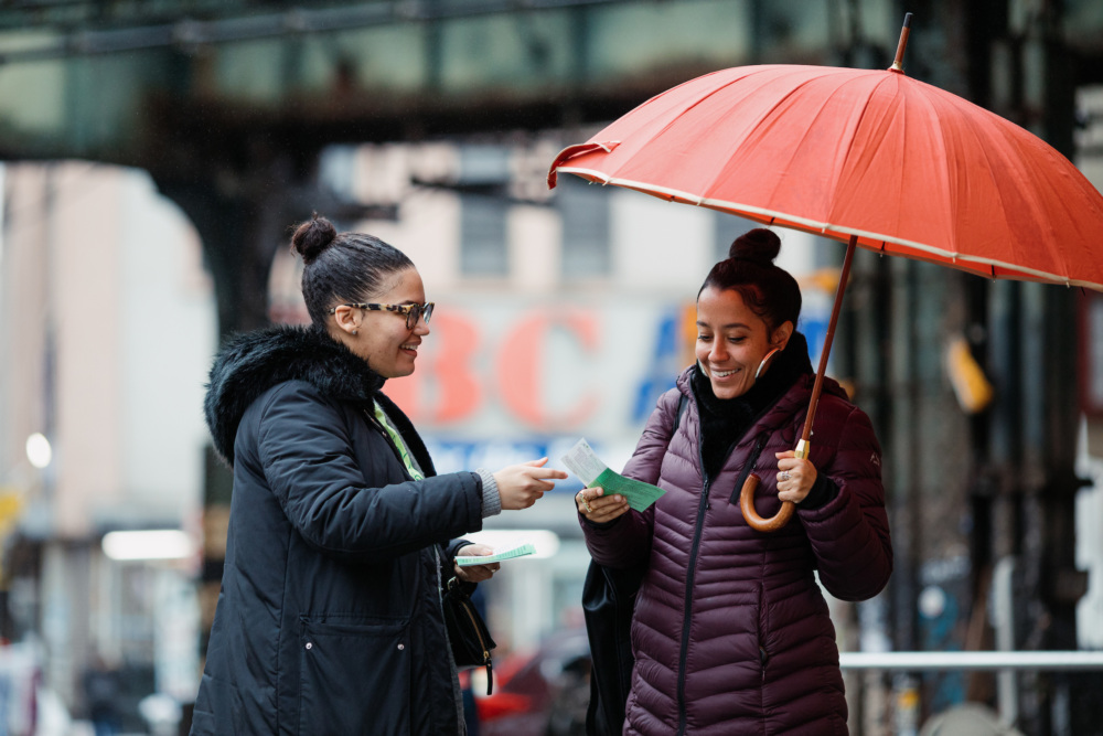 Two women sharing a laugh under a red umbrella on a rainy city street, one holding a pamphlet and the other carrying a white bag.