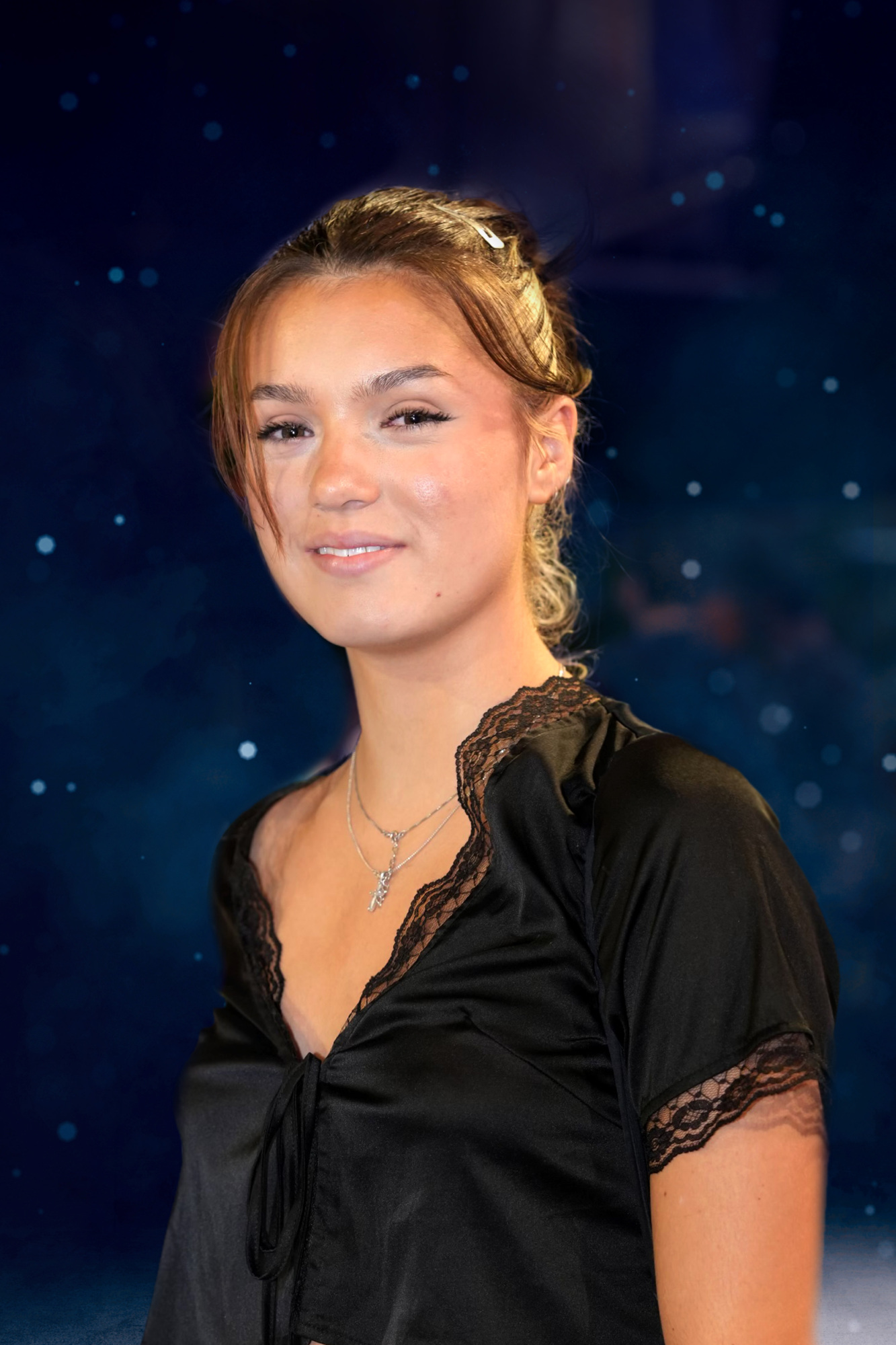 Young woman with a relaxed smile, wearing a black lace-trimmed blouse, with her hair pulled back. a blurred blue background with sparkles enhances the portrait.
