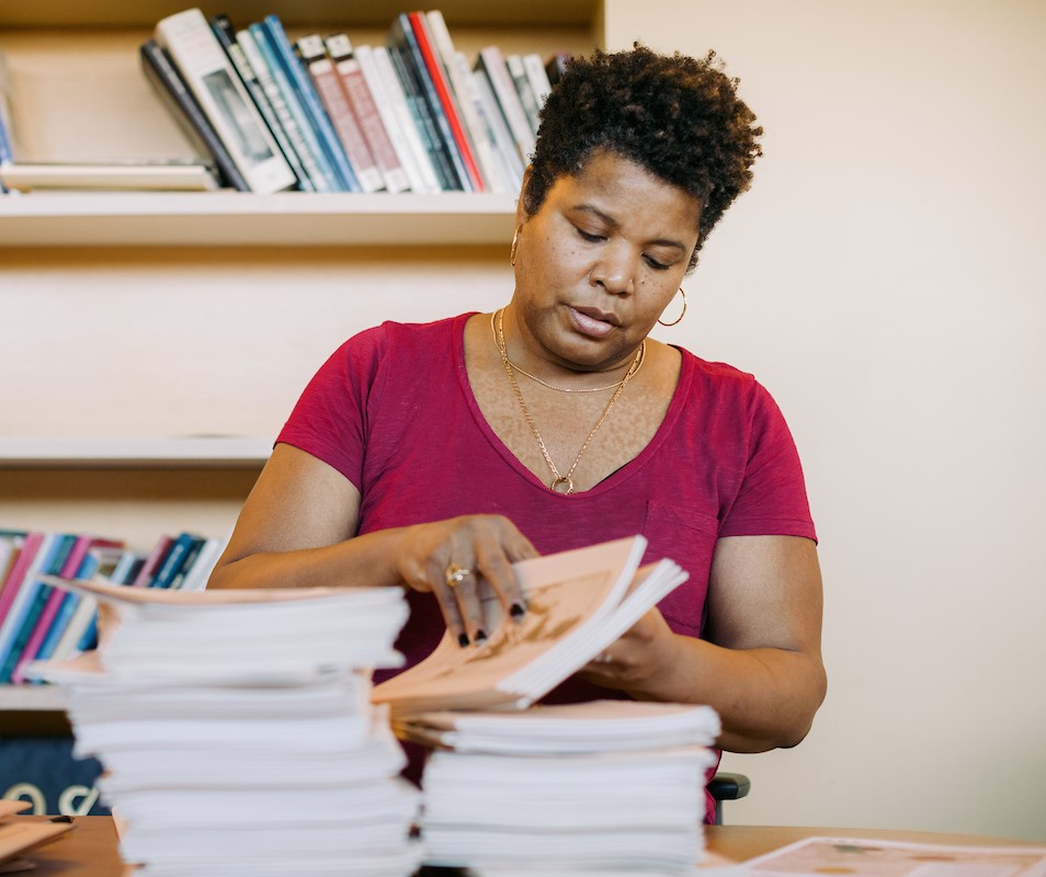 A woman in a red shirt organizes a stack of papers at a desk surrounded by books.