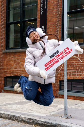 Woman joyfully jumping with a sign promoting brooklyn gives on giving tuesday.