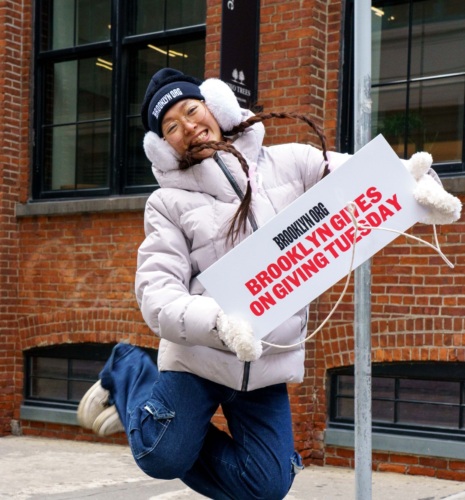 Woman joyfully jumping with a sign promoting brooklyn gives on giving tuesday.