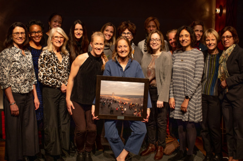 Group of fifteen women smiling at a gala event, one of them holding a framed photograph. they are standing on a stage with a red curtain backdrop.
