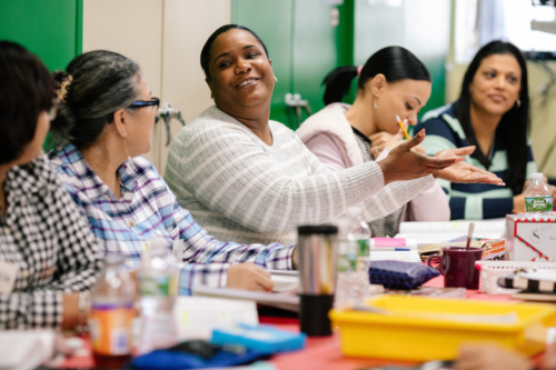 A group of people sit around a table in discussion, with notebooks, water bottles, and stationery on the table. One person in the center is speaking and gesturing with their hands.