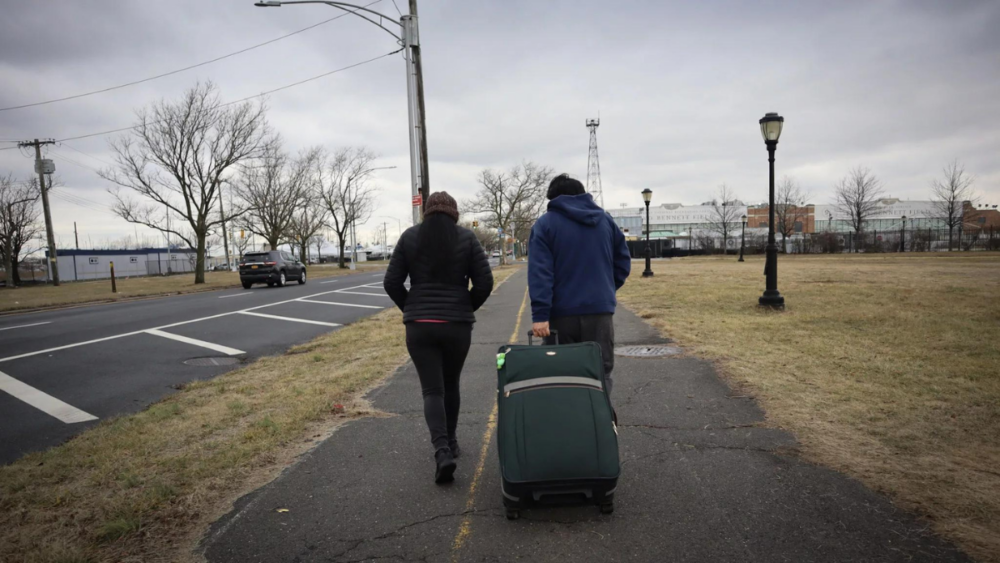 Two people walking along a sidewalk near a road, one pulling a green suitcase, in an overcast urban setting.
