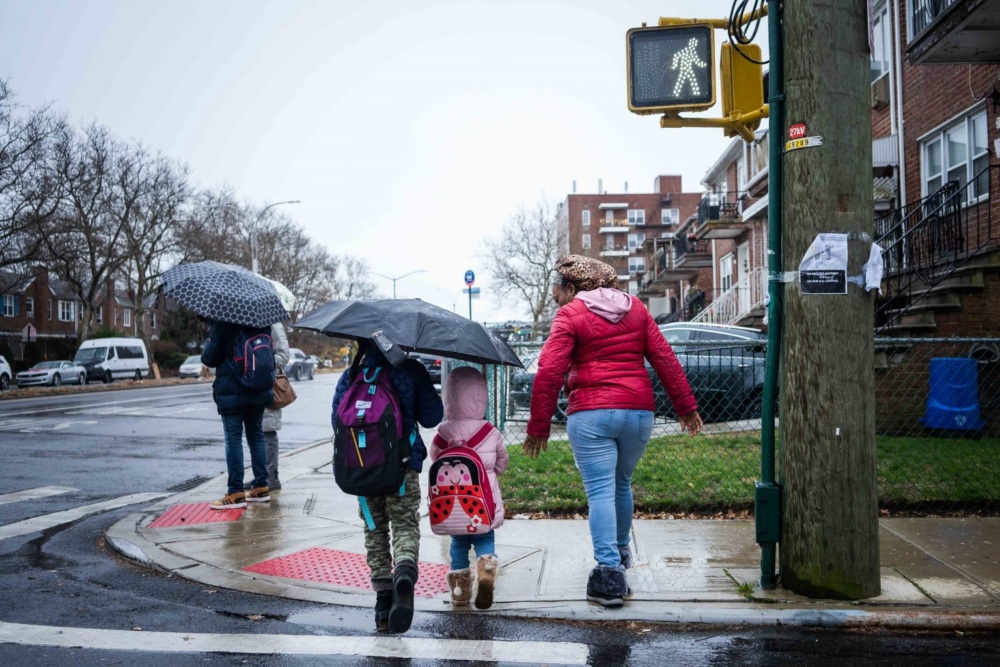 A group of people, including children with backpacks and a woman with an umbrella, cross a rainy street at a crosswalk.